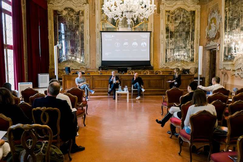 In una sala con ricche decorazioni barocche, quattro persone parlano a un folto pubblico seduto