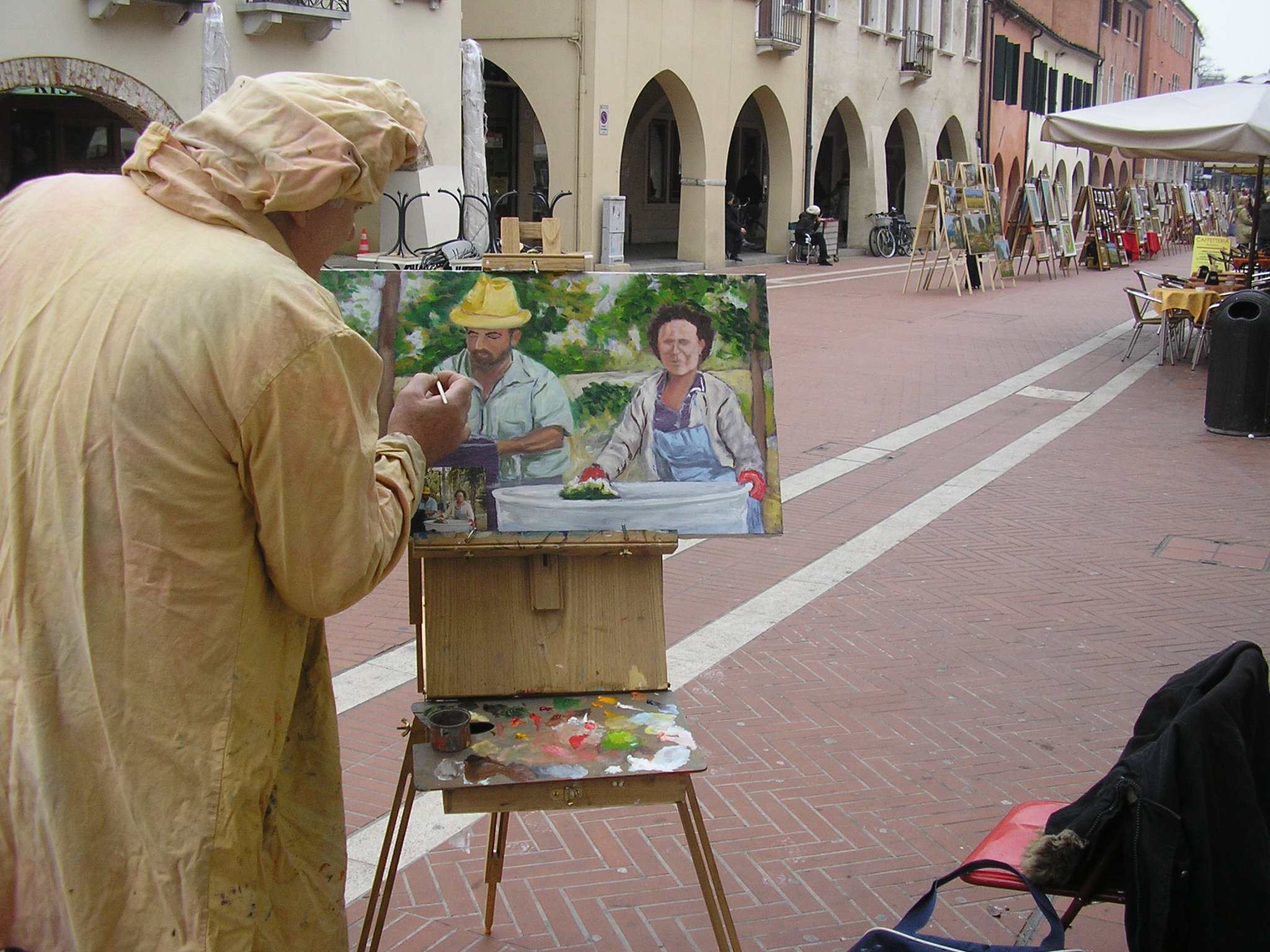 Pittore visto di spalle dipinge opera su un cavalletto posizionato all'aperto in una via cittadina