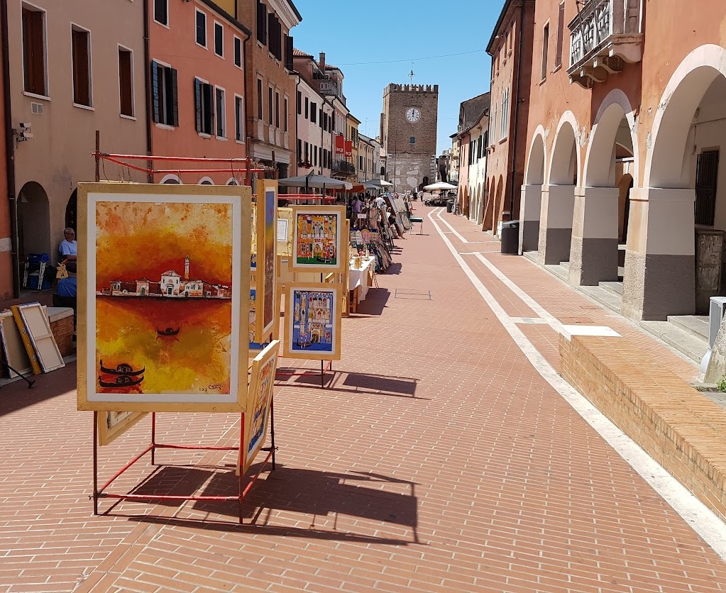 Quadri esposti su cavalletti in una via cittadina porticata. Sullo sfondo una torre con orologio