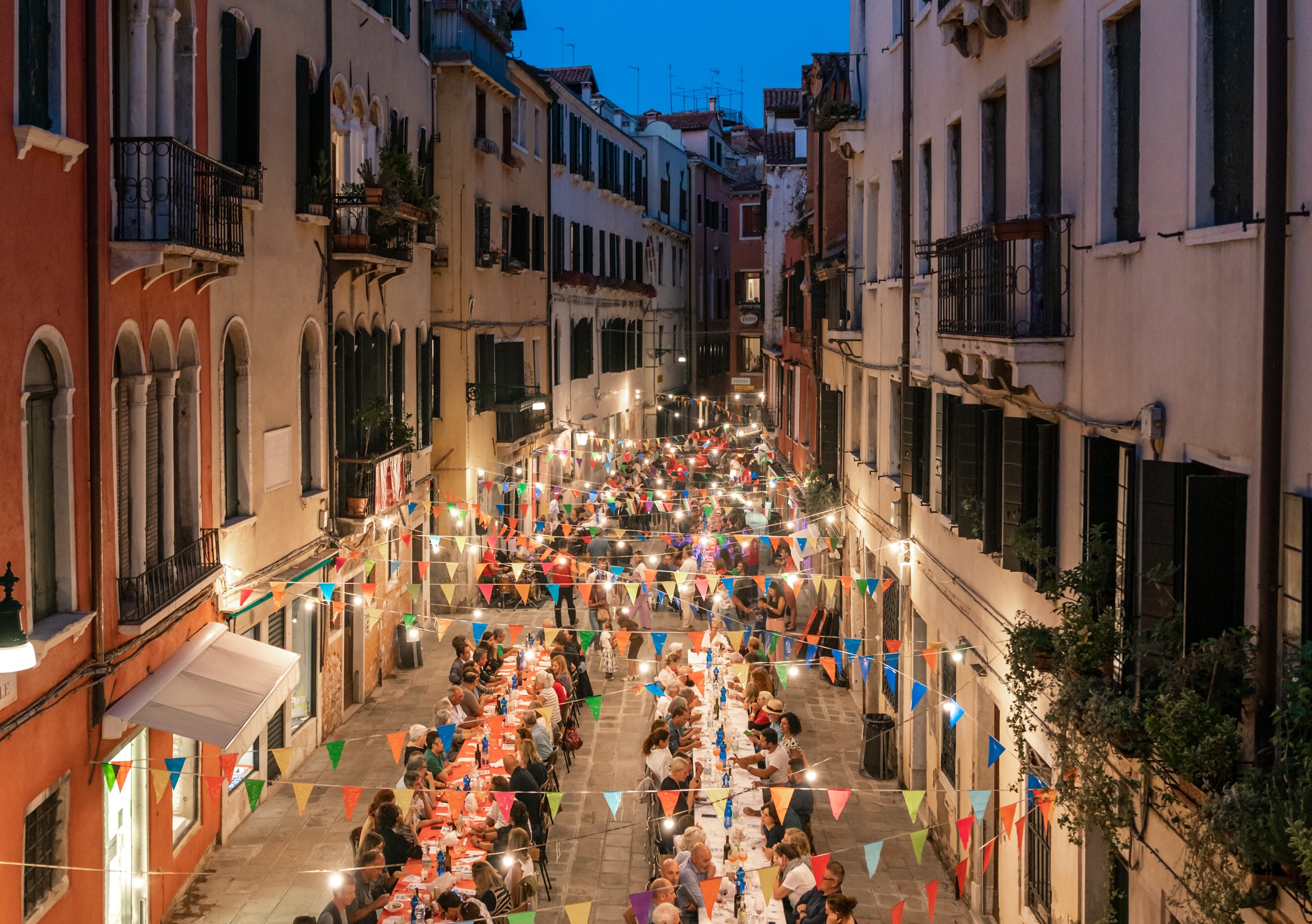Lunghe tavolate all'aperto organizzate in una strada veneziana di sera