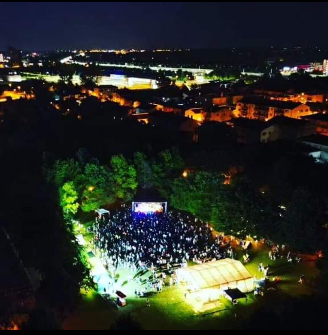 Palco illuminato di notte con, di fronte, un folto pubblcio; sullo sfondo le luci notturne di una città