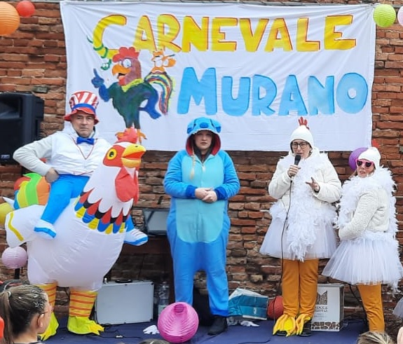 Tre persone vestite da gallina e una da cane su di un palco. Sullo sfondo uno striscione con scritto "Carnevale di Murano"