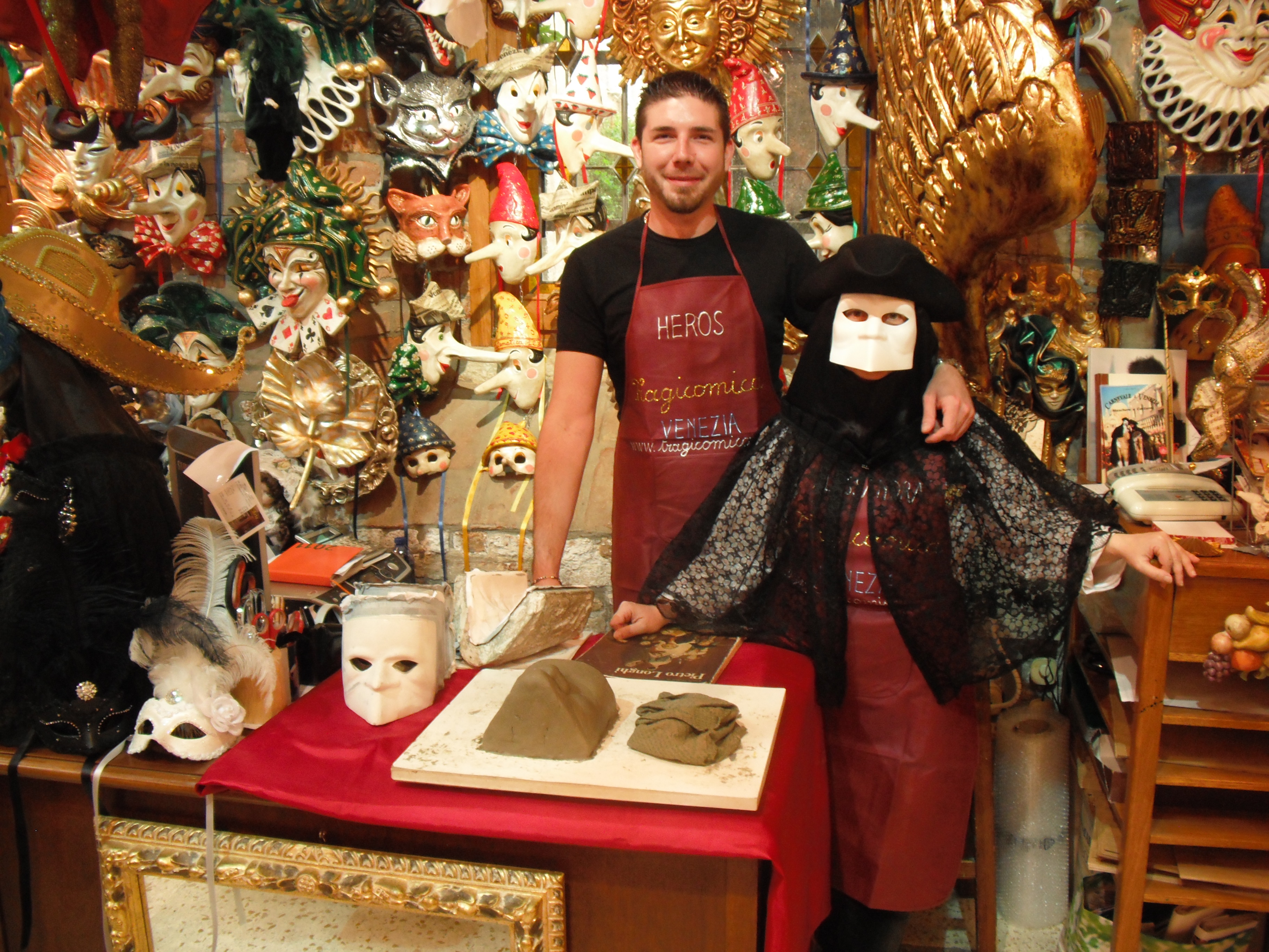 Un uomo e una persona che indossa tipica maschera veneziana posano davanti a una parete ricoperta di maschere di ogni tipo