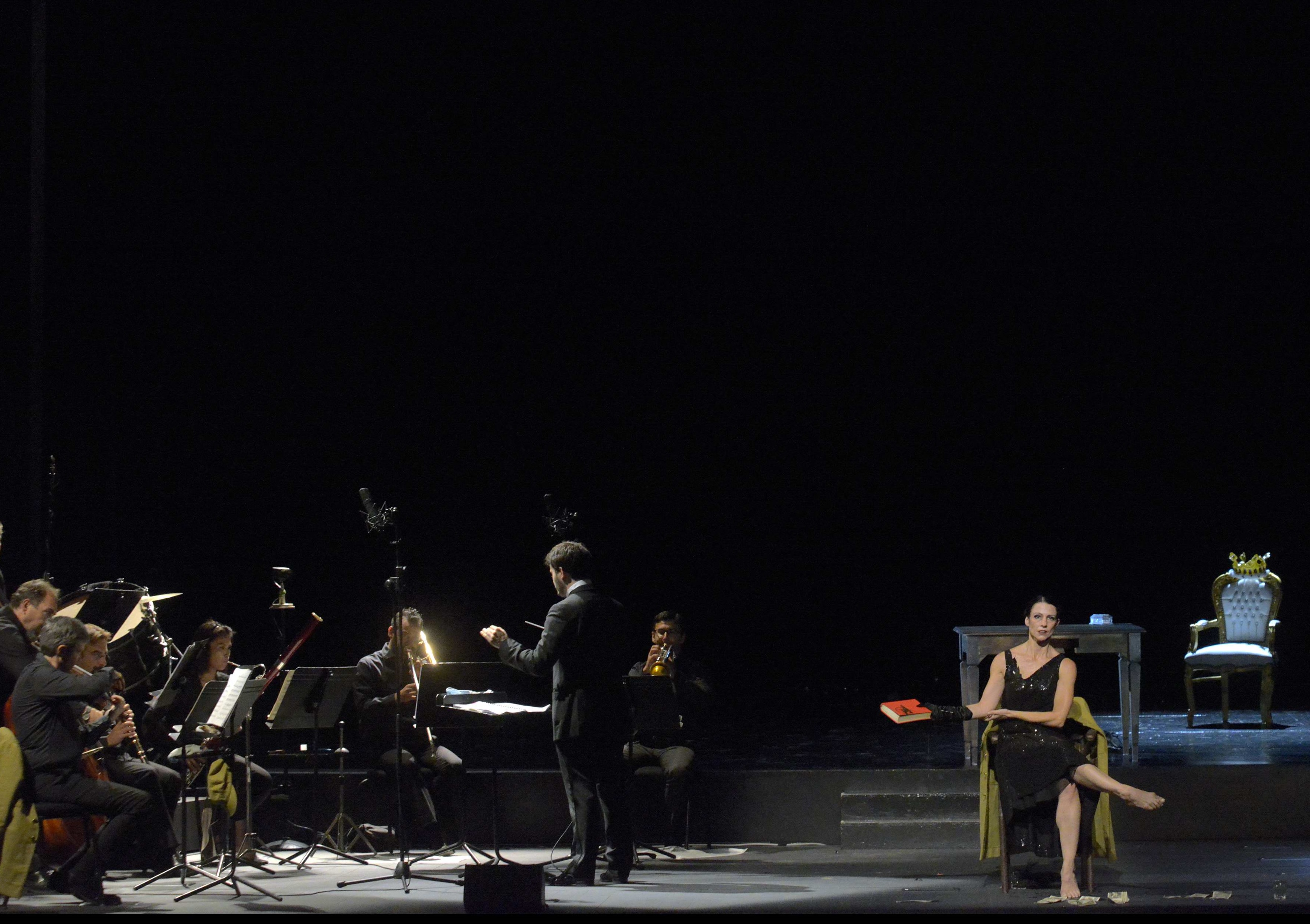 A sinistra orchestra, a destra donna che regge un libro, seduta su una sedia sopra al palco