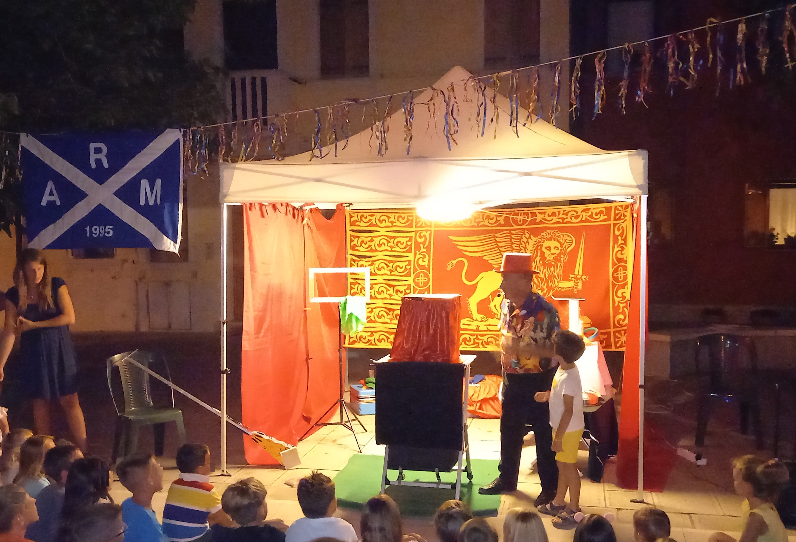 Pubblico di bambini guarda un mago sul palco accompagnato da un altro bambino