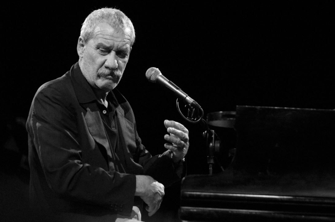 Primo piano di uomo canuto e coi baffi (Paolo Conte), che regge un microfono