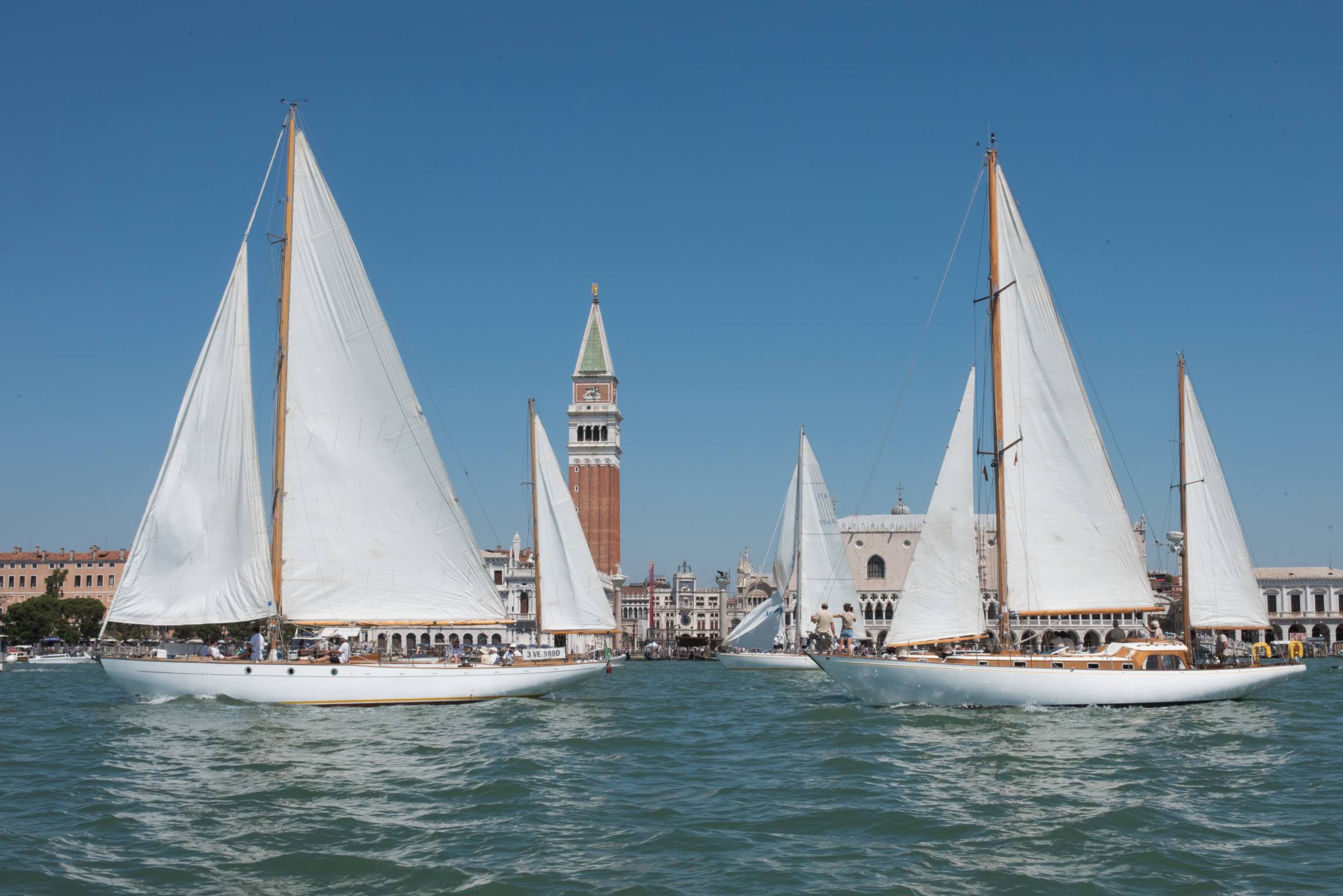 Due barche a vela in primo piano e una terza sullo sfondo transitano di fronte a piazza S. Marco. Dalle vele svetta il campanile
