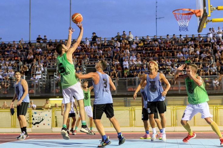 Dei giocatori (8) si affrontano durante una partita di basket. Uno sta tirando in sospensione. Sullo sfondo spalti gremiti.