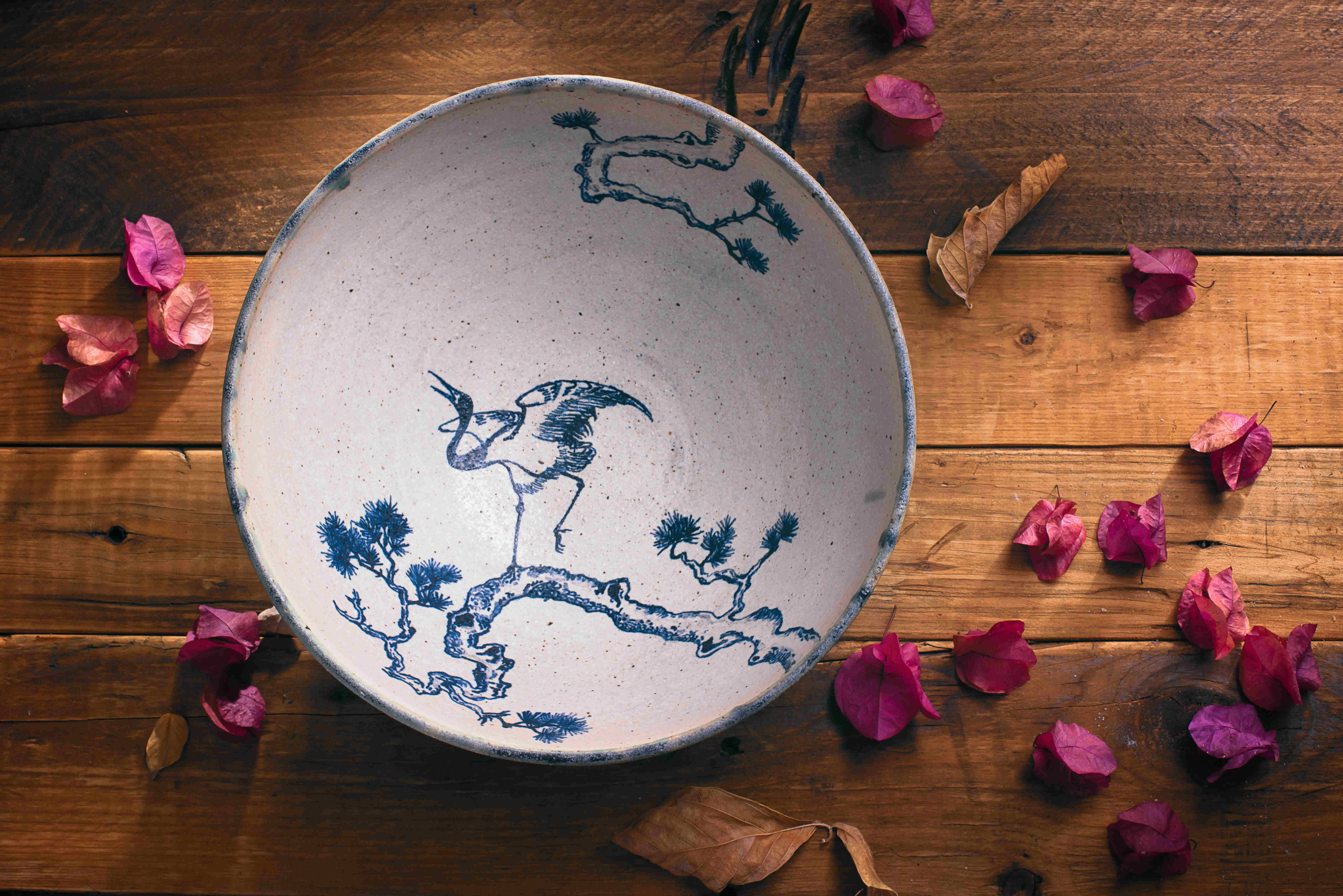 Piatto in ceramica che raffigura un airone, appoggiato su un piano di assi di legno cosparso di petali